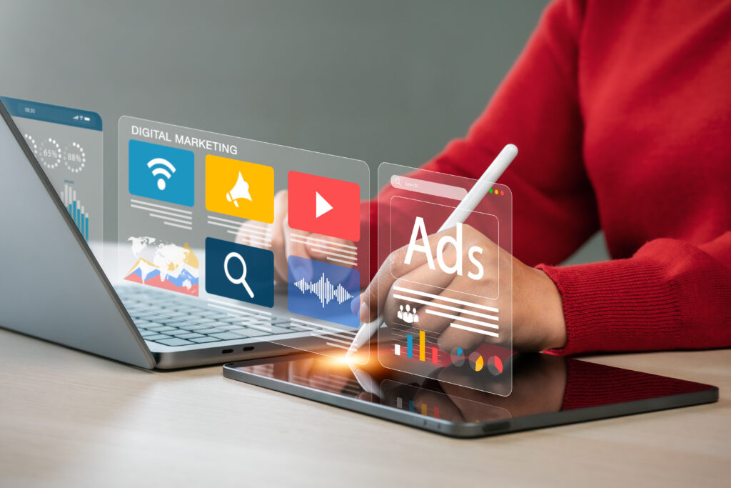 Frau arbeitet mit iPad und Laptop, darüber schweben Kacheln mit Symbolen und Anzeigen zu digitalem Marketing und Werbung